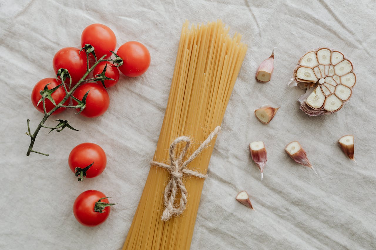 Flat lay of cherry tomatoes, garlic, and uncooked spaghetti on a neutral background. Ideal for food photography.