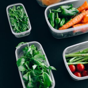Assorted fresh vegetables including arugula, carrots, and tomatoes in plastic containers against a black background.