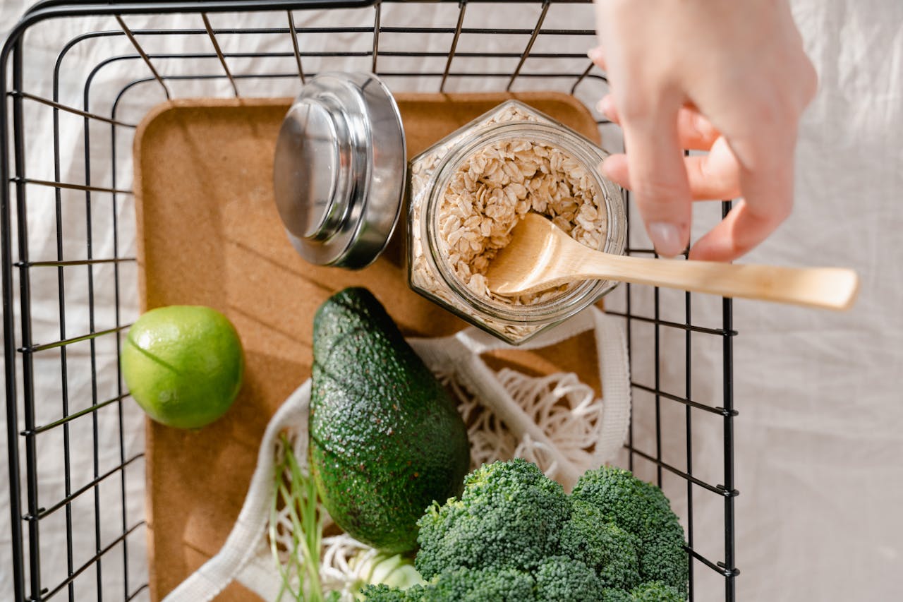 A metal basket with fresh vegetables, avocado, lime, and oats arranged in a flat lay style.