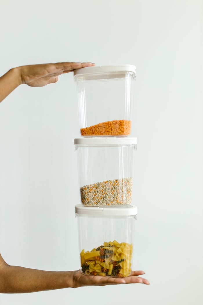 Hands holding stacked plastic containers with various dry foods against a neutral background.