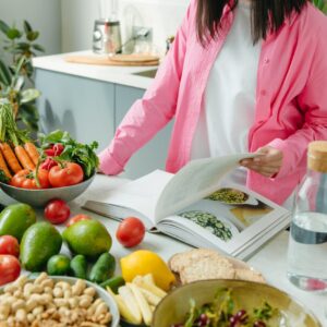 A person browsing a cookbook surrounded by fresh vegetables in a bright kitchen setting.